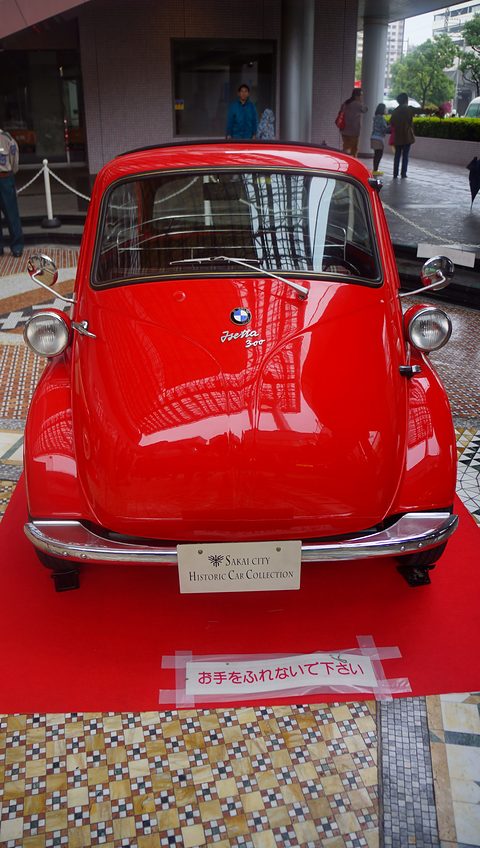 Red bubble car on a red carpet seen straight from the front, a shopping centre behind. Opens a larger view