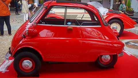 Red bubble car in side view on a red carpet, visitors behind. Opens a larger view