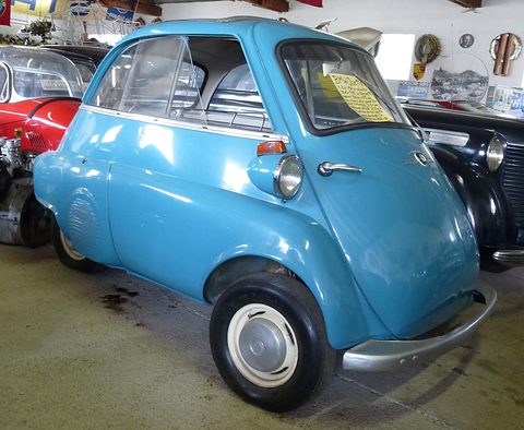 Bright blue bubble car on a museum floor among other cars, seen from the front right. Opens a larger view