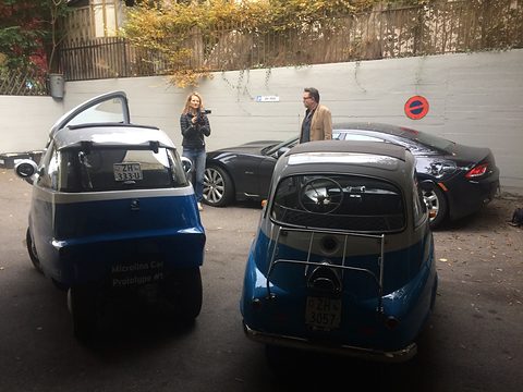 Two blue and white bubble cars parked side by side seen from behind, the Isetta with a luggage rack, two people talking beyond. Opens a larger view