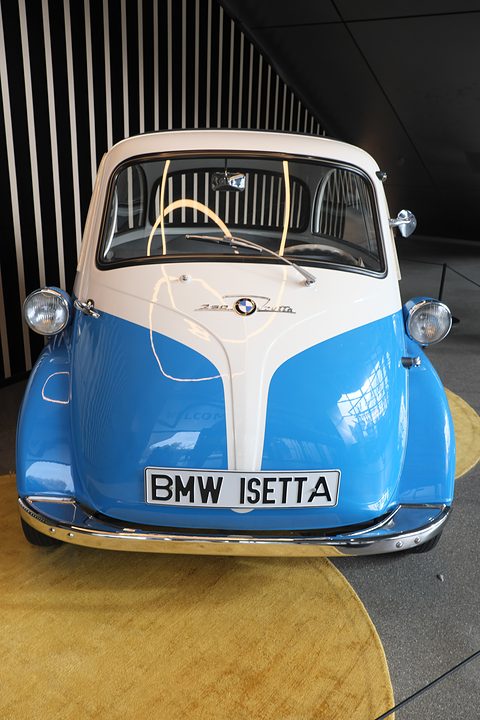 Blue and white bubble car seen straight from the front with a BMW Isetta plate, striped wall behind. Opens a larger view