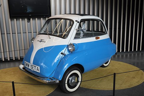 Blue and white bubble car with whitewall tyres on a yellow round carpet, seen from the front left. Opens a larger view