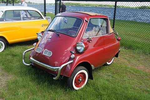 Dark red bubble car with a fabric roof on grass beside a fence, a yellow car behind. Opens a larger view