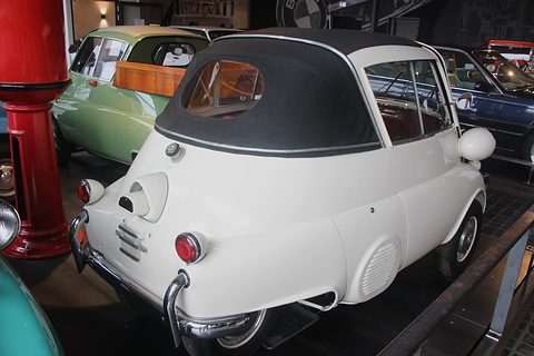 White bubble car with a black fabric roof seen from behind in a museum hall. Opens a larger view
