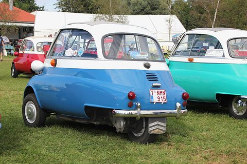 Blue and white bubble car seen from the rear on grass, its single rear wheel visible, other Isettas around