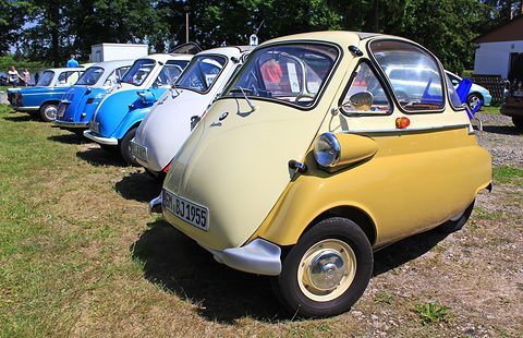 Row of bubble cars on grass, a pale yellow one at the front seen from the front right. Opens a larger view