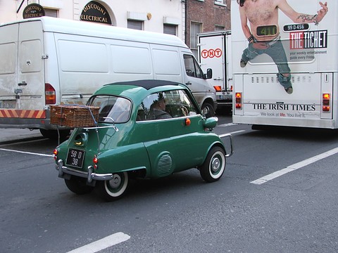 Green bubble car seen from the side on a street, a white van and a bus behind. Opens a larger view
