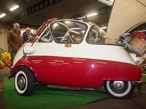 Red and white bubble car with whitewall tyres on green carpet in a hall, two visitors behind. Opens a larger view