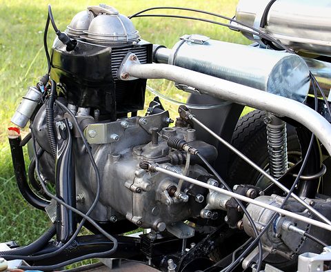 Bare engine with cooling fins, carburettor and gearbox resting on grass. Opens a larger view