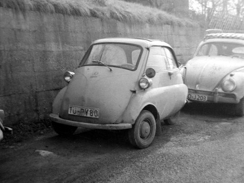 Worn bubble car parked against a rough stone wall next to a Beetle, in black and white. Opens a larger view