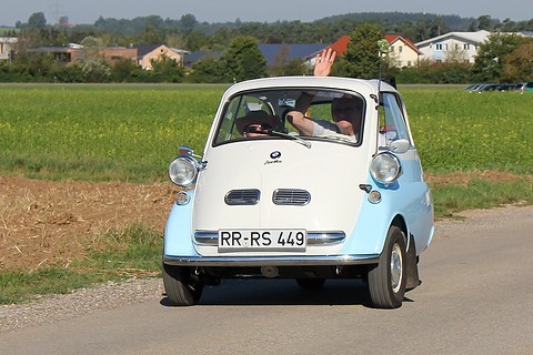 Blue and white bubble car driving towards the camera on a country road, two people inside. Opens a larger view