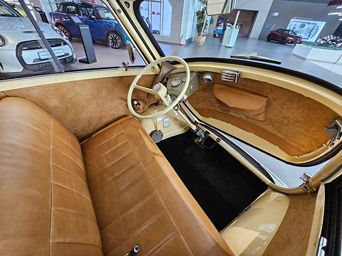 Tan leather bench seat and a cream steering wheel inside an open bubble car, showroom behind. Opens a larger view