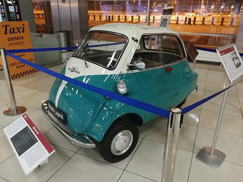 Teal and white bubble car inside a blue rope barrier on a tiled airport floor, a taxi sign behind. Opens a larger view