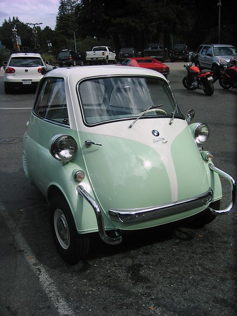 Mint green and white bubble car seen from the front on tarmac, motorcycles behind. Opens a larger view
