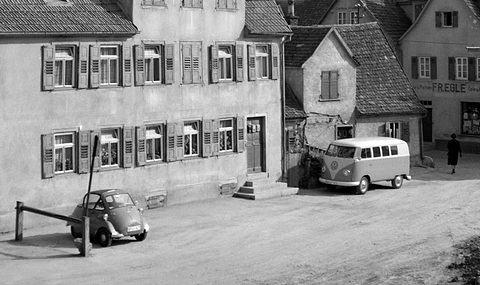 Old half-timbered houses around a sloping yard with a small bubble car at left and a VW bus at right. Opens a larger view