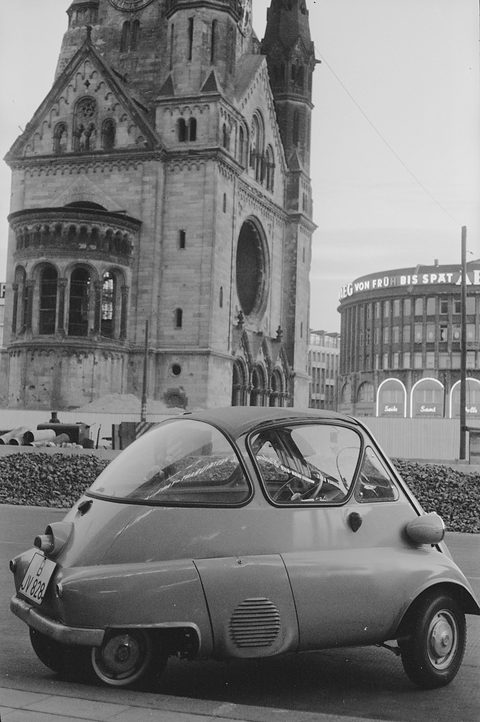 Bubble car parked at a kerb in front of the war-damaged church tower and a round modern building. Opens a larger view