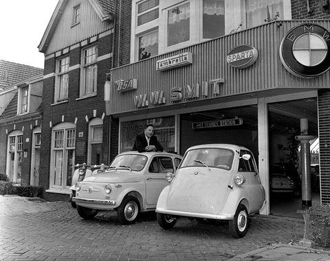 Two small cars parked on cobbles in front of a dealer’s garage with BMW and Lambretta signs, a man leaning on the Fiat