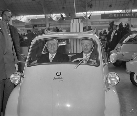 Front of a pale bubble car with an Isetta 300 badge, two men in suits behind its windscreen, onlookers around it. Opens a larger view