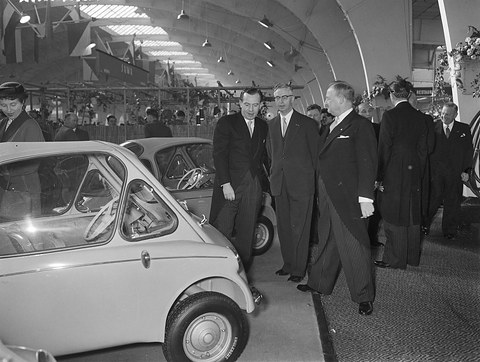 Men in dark coats walking along a row of exhibits in a hall, an Isetta in the left foreground