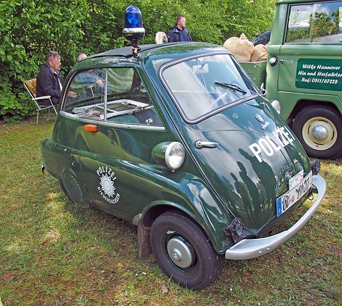 Dark green bubble car with Polizei lettering and a blue beacon, parked on grass under trees. Opens a larger view