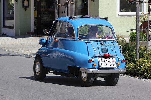 Blue bubble car seen from behind on a road with a chrome luggage rack, a house behind. Opens a larger view