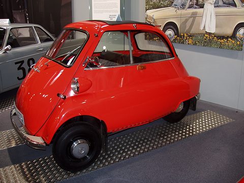 Red bubble car on a ribbed metal stand, a racing car and posters behind. Opens a larger view