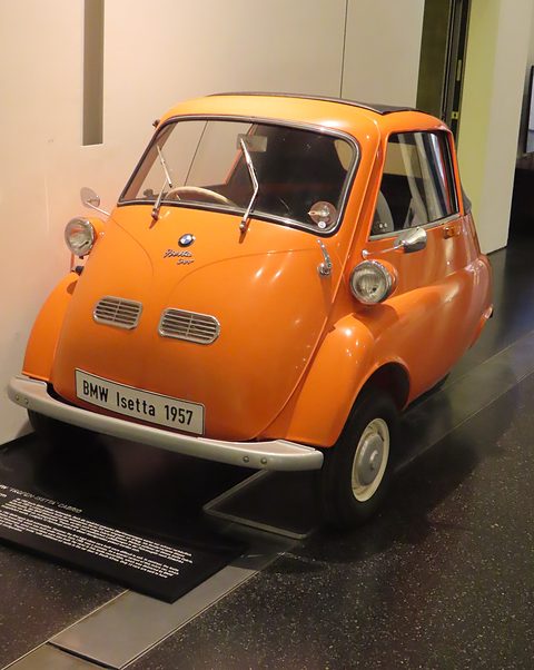 Orange bubble car with a dark fabric roof on a black floor in a white museum room. Opens a larger view