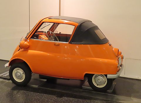 Orange bubble car in side view with a black folding roof, against a white wall. Opens a larger view
