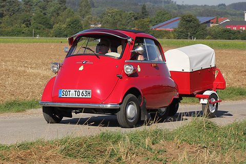Red bubble car towing a red and white trailer along a road beside a field. Opens a larger view