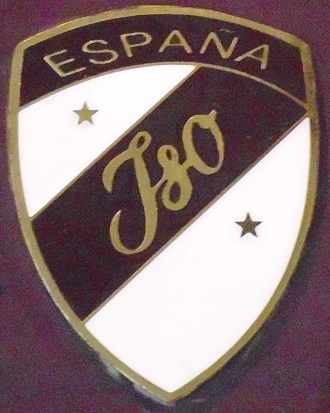 Shield-shaped enamel badge, maroon and white, with ESPAÑA across the top, two stars and a gold Iso script. Opens a larger view