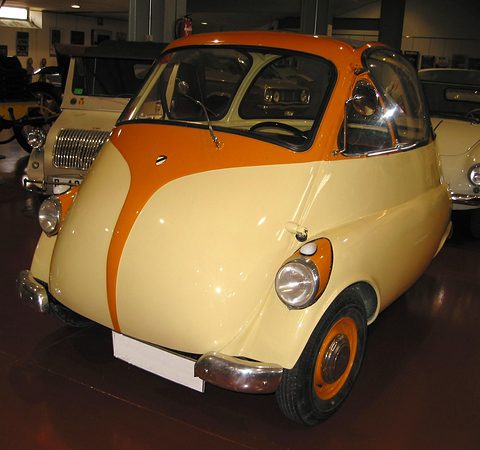 Cream and orange two-tone bubble car in a museum hall, an orange stripe sweeping down its nose between low headlamps. Opens a larger view