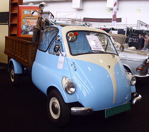 Pale blue Isetta-cab pick-up seen from the front right, its wooden load bed behind the cab, other classics behind. Opens a larger view