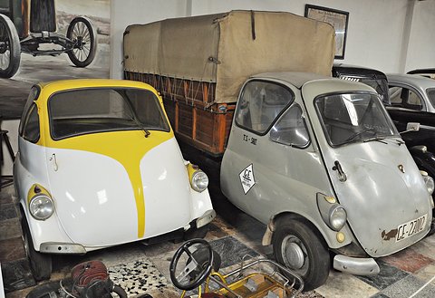 A grey Isetta-cab truck with a canvas tilt beside a white and yellow bubble car in a crowded museum room. Opens a larger view