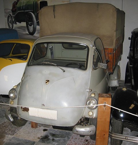 Grey Isetta-cab truck with a canvas-covered load bed behind a museum rope, a yellow car beside it. Opens a larger view