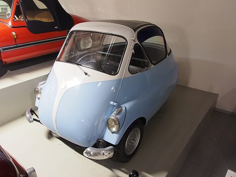 Pale blue and white bubble car on a grey museum plinth, seen from the front left, an orange open car parked above it. Opens a larger view