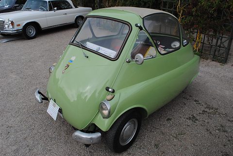 Pale green bubble car on gravel from the front right, small headlamps low on the wings, a white saloon behind. Opens a larger view