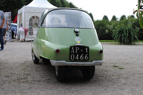 Rear of a pale green bubble car with a black Italian number plate, two narrow-set rear wheels and a white marquee behind. Opens a larger view