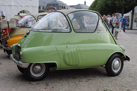 Side view of a pale green egg-shaped car with a slotted panel over the engine and a large wraparound window, visitors and tents behind. Opens a larger view