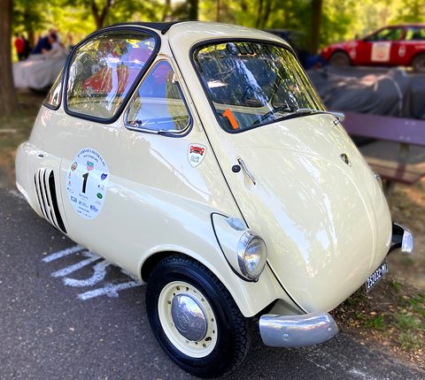 Cream bubble car parked under trees, a round rally plate with the number 1 on its side, slotted engine cover behind. Opens a larger view