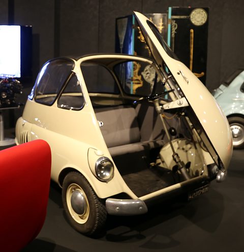 Cream bubble car with its front door raised and the steering column swung out with it, showing the bench seat, in a dark gallery.
