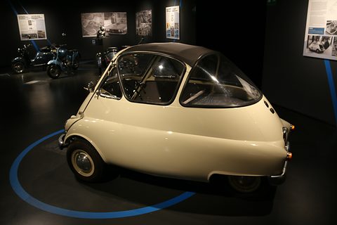 Cream bubble car with a dark roof on a black museum floor marked with a blue circle, display panels behind. Opens a larger view