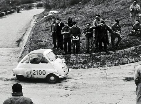 Black-and-white photograph of a small egg-shaped car numbered 2100 cornering on a hairpin, men watching from the bank. Opens a larger view