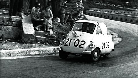 Black-and-white photograph of a small white bubble car numbered 2102 leaning through a bend past spectators sitting on a stone wall.