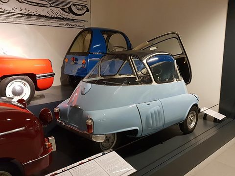 Pale blue egg-shaped car from behind and to the side, a slotted engine cover on its flank, a red car’s tail at left. Opens a larger view
