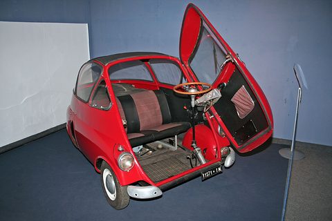 Red bubble car with its front door swung wide, the steering wheel on the door and a bench seat inside. Opens a larger view