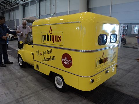 Yellow three-quarter rear view of a box van with an Isetta cab, signwritten for a bottled-gas company, in an exhibition hall. Opens a larger view
