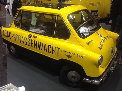 Yellow and black small car lettered ADAC-STRASSENWACHT along its side, on a show stand.