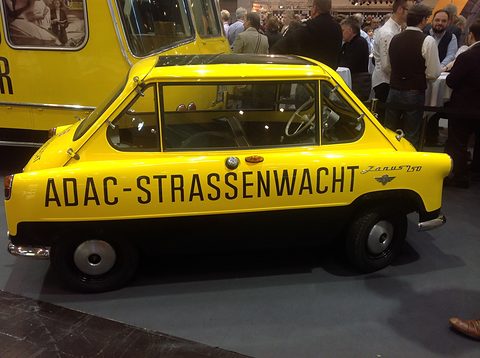 Yellow small car in side view with ADAC-STRASSENWACHT in black capitals, visitors crowded behind. Opens a larger view