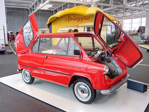 Red small car in side view with front and rear doors swung up, a yellow beach-hut display behind. Opens a larger view