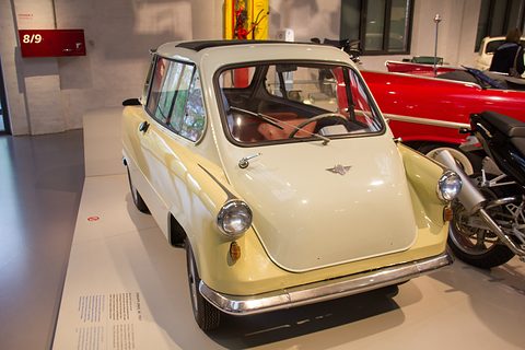 Cream and white small car with a winged badge on its nose door, on a pale plinth beside a red car. Opens a larger view